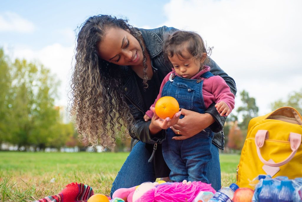 A loving mother with a child in a park holding an orange, enjoying a sunny day outdoors