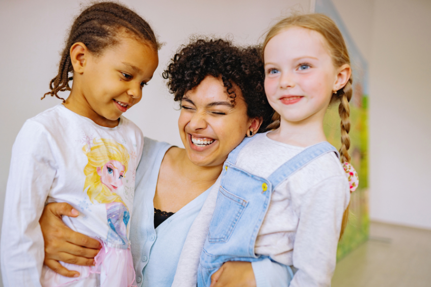 A smiling teacher laughing while embracing two preschoolers, one on each side, in a bright indoor classroom space 